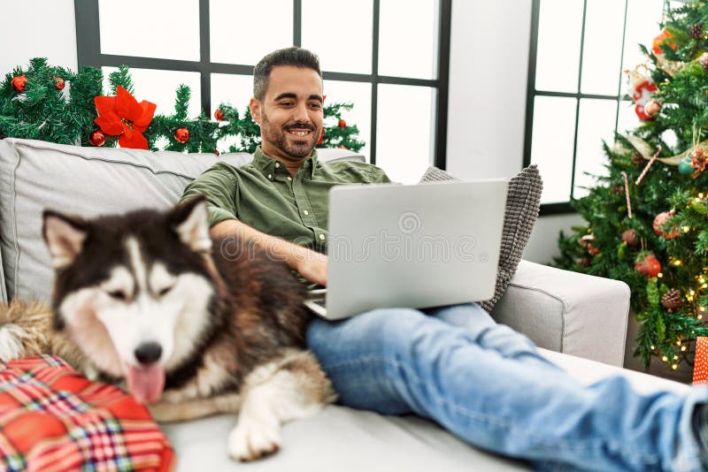 Young hispanic man using laptop sitting on sofa with dog by christmas tree at home