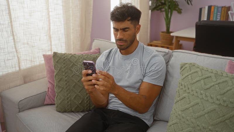 Young hispanic man using smartphone while sitting on a couch in a cozy living room stock photo