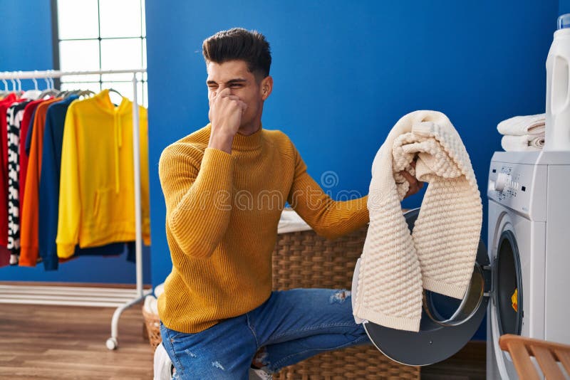 Young Hispanic Man Washing Clothes Smelling Dirty Clothes at Laundry
