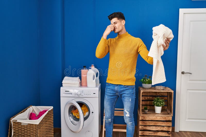 Young Hispanic Man Washing Clothes Smelling Dirty Clothes at Laundry