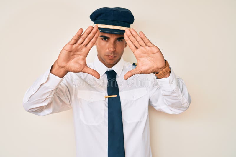 Young hispanic man wearing airplane pilot uniform doing frame using hands palms and fingers, camera perspective stock photography