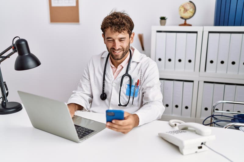 Young hispanic man wearing doctor uniform using smartphone at clinic stock photography