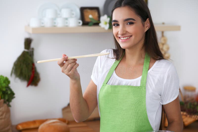 Young Hispanic Woman In A Green Apron Cooking In The Kitchen. Housewife Holding Wooden Spoon While Smiling.