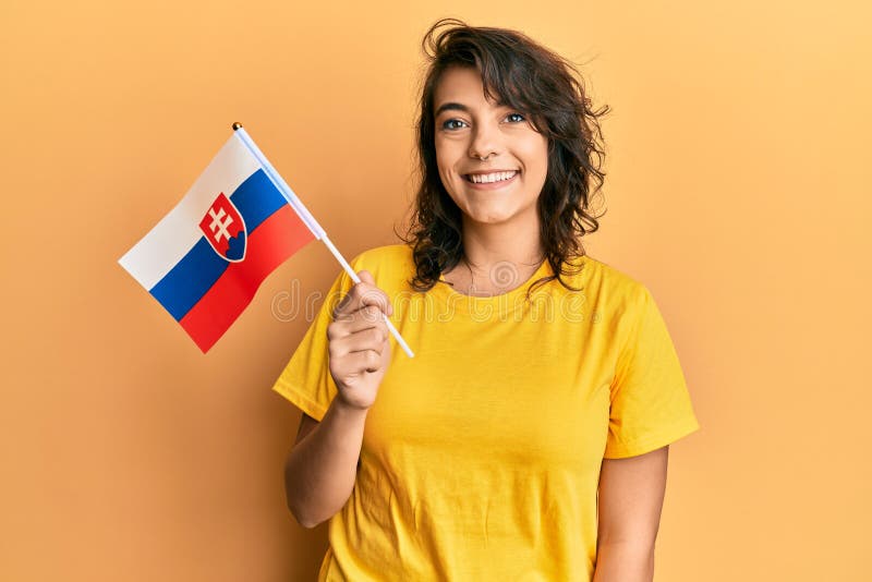 Mladá hispánka drží slovenskú vlajku vyzerá pozitívne a šťastne stojaca a usmievavá so sebavedomým úsmevom ukazujúcim zuby
