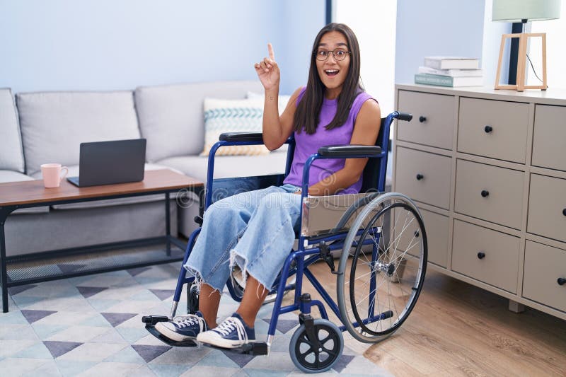 Young hispanic woman sitting on wheelchair at home pointing finger up with successful idea