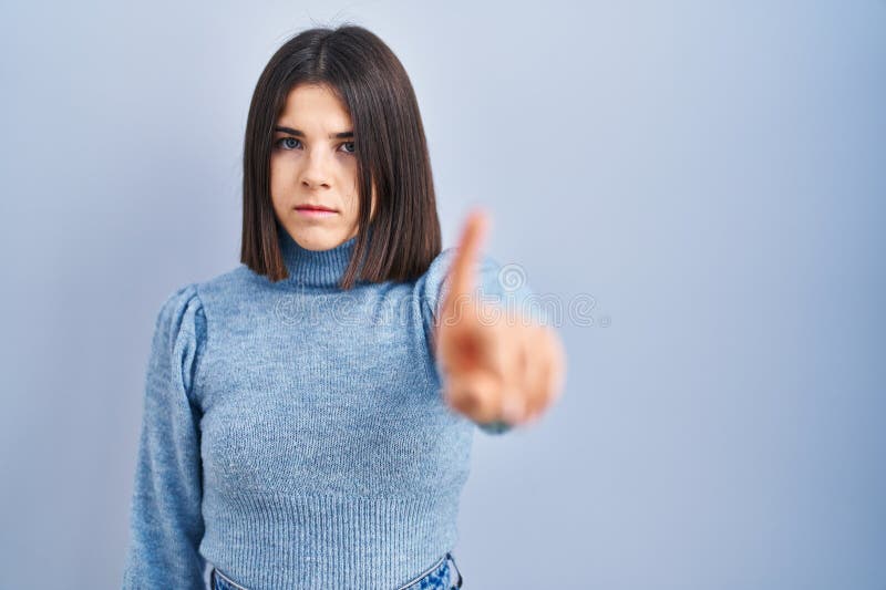 Young hispanic woman standing over blue background pointing with finger up and angry expression, showing no gesture