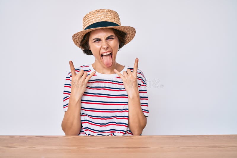 Young hispanic woman wearing casual clothes and hat sitting on the table shouting with crazy expression doing rock symbol with