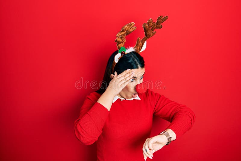 Young hispanic woman wearing deer christmas hat and red nose looking at the watch time worried, afraid of getting late