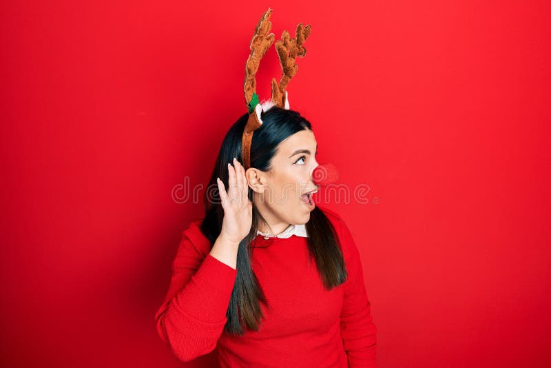 Young hispanic woman wearing deer christmas hat and red nose smiling with hand over ear listening an hearing to rumor or gossip