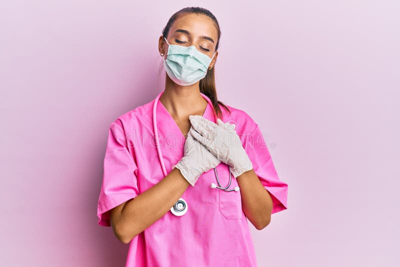 Young hispanic woman wearing doctor uniform and medical mask smiling with hands on chest, eyes closed with grateful gesture on