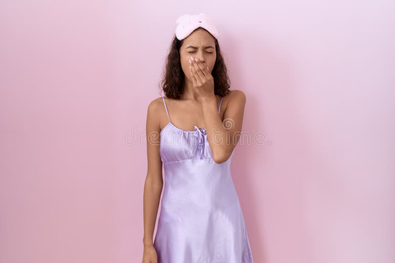 Young hispanic woman wearing sleep mask and nightgown bored yawning tired covering mouth with hand