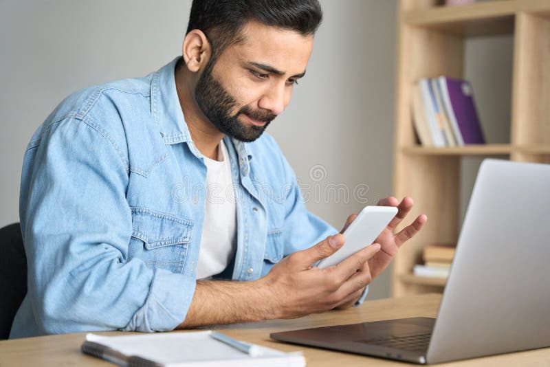 Young indian businessman works from home office using cellphone and laptop.