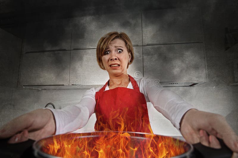 Young inexperienced home cook woman in panic with apron holding pot burning in flames with in panic