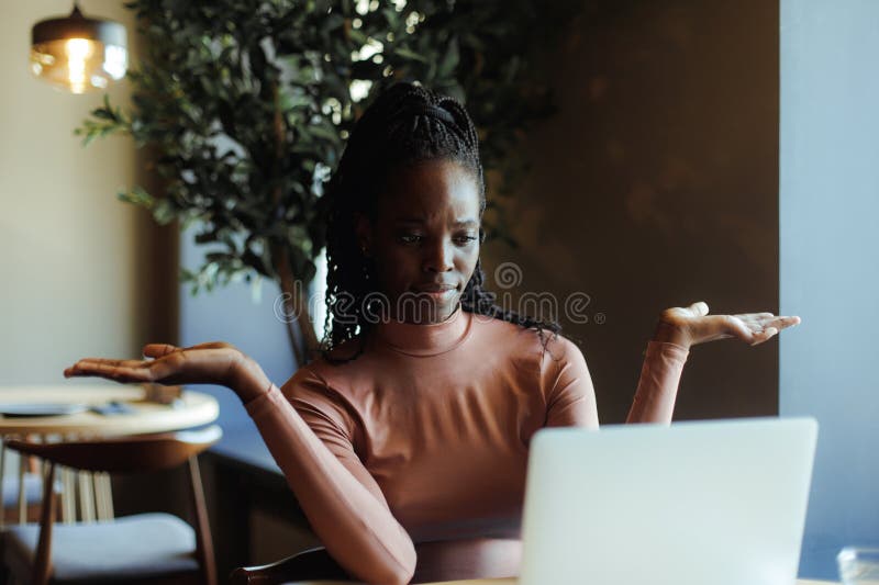 Young irritated African-American woman sit at table, looking at screen of laptop, raising arms in questioning gesture. royalty free stock photos
