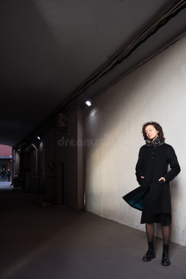 The young lady in black stylish coat with black scarf enjoying the sun rays at sunset in the white tunnel in the cold autumn day and black shadow on the white wall. The young lady in black stylish coat with black scarf enjoying the sun rays at sunset in the white tunnel in the cold autumn day and black shadow on the white wall