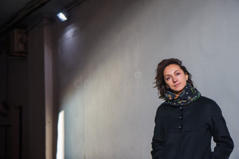 The young lady in black stylish coat with black scarf enjoying the sun rays at sunset in the white tunnel in the cold autumn day and black shadow on the white wall. The young lady in black stylish coat with black scarf enjoying the sun rays at sunset in the white tunnel in the cold autumn day and black shadow on the white wall