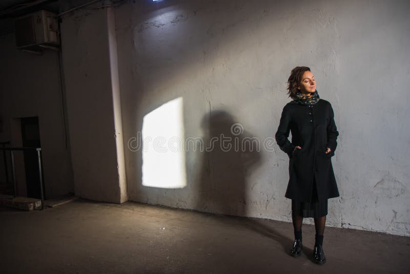 The young lady in black stylish coat with black scarf enjoying the sun rays at sunset in the white tunnel in the cold autumn day and black shadow on the white wall. The young lady in black stylish coat with black scarf enjoying the sun rays at sunset in the white tunnel in the cold autumn day and black shadow on the white wall