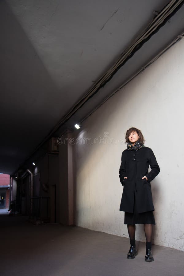 The young lady in black stylish coat with black scarf enjoying the sun rays at sunset in the white tunnel in the cold autumn day and black shadow on the white wall. The young lady in black stylish coat with black scarf enjoying the sun rays at sunset in the white tunnel in the cold autumn day and black shadow on the white wall