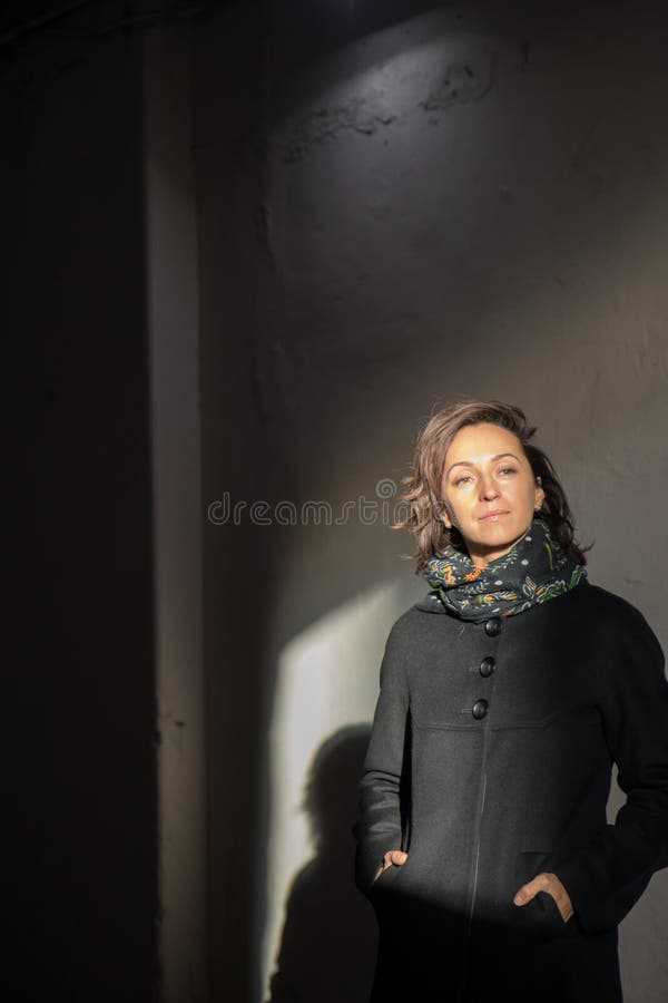 The young lady in black stylish coat with black scarf enjoying the sun rays at sunset in the white tunnel in the cold autumn day and black shadow on the white wall. The young lady in black stylish coat with black scarf enjoying the sun rays at sunset in the white tunnel in the cold autumn day and black shadow on the white wall