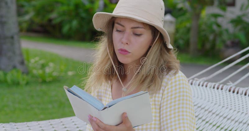 Young lady in hat with book in hammock. Charming happy woman resting in hammock and reading book with exciting plot at