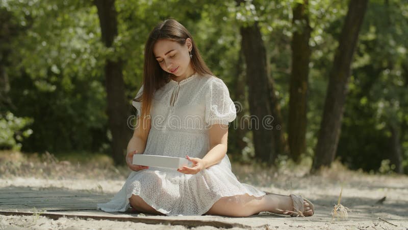 A young lady sits on the sand in a summer forest. A girl in a white dress holds a box with a birthday gift in her hands