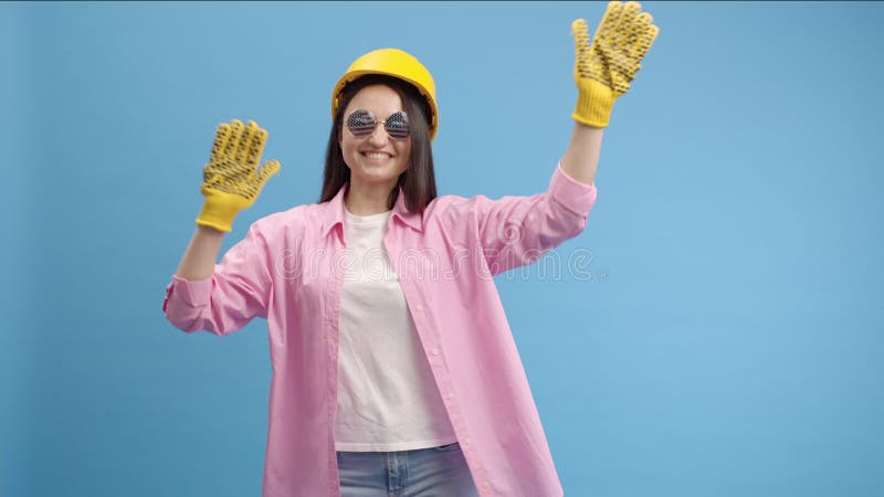 Young lady worker in yellow hard hat, American glasses and gloves. Dancing in studio on blue background and smiling