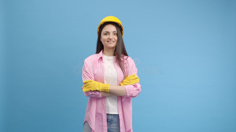 Young lady worker in yellow hard hat and gloves. Posing in studio on blue background and smiling