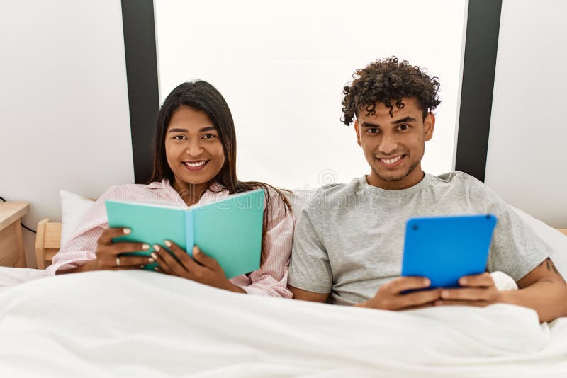 Young Latin Couple Reading Book Using Touchpad Sitting on the Bed at Bedroom Stock Photo Image