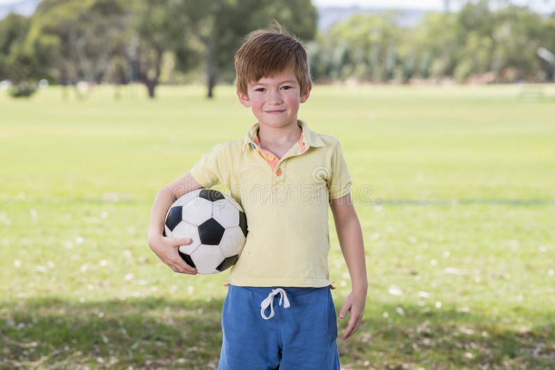 Young little kid 7 or 8 years old enjoying happy playing football soccer at grass city park field posing smiling proud standing ho