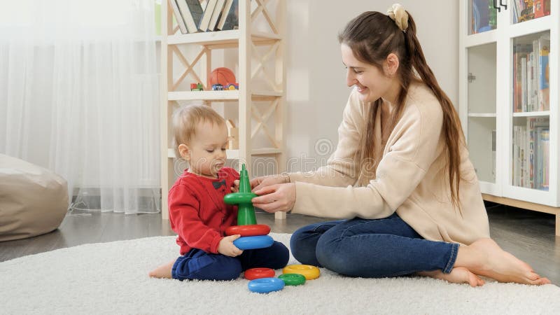 Young loving mother helping her baby son assembling colorful toy tower or pyramid