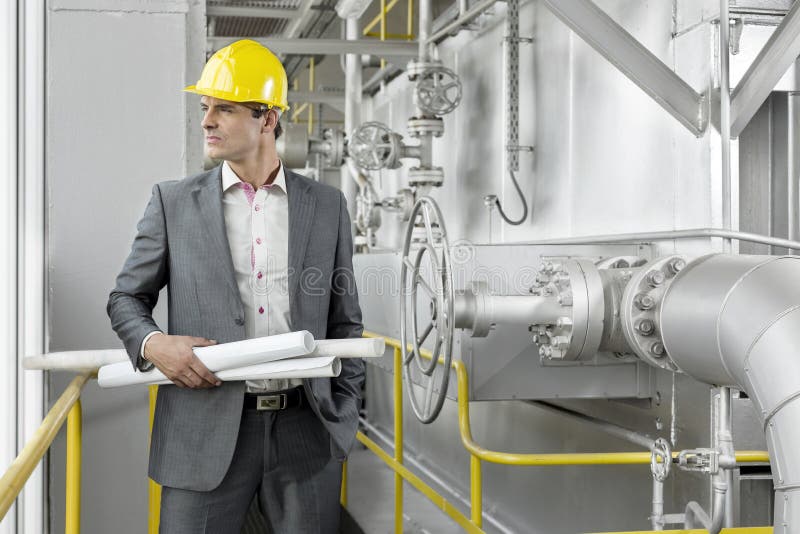 Young male architect holding rolled up blueprints by industrial machinery