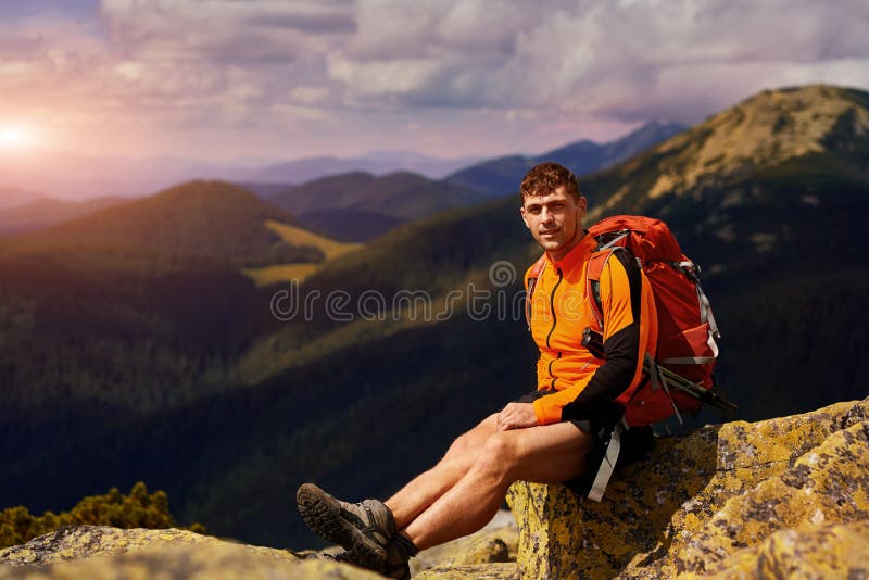Young male hiker with backpack relaxing on top of a mountain during calm summer sunset