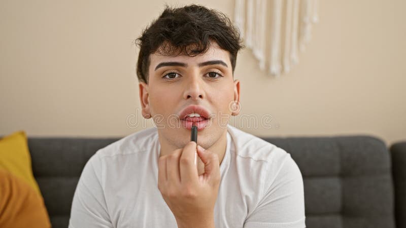 A young man applies lipstick while seated indoors, portraying a cozy, casual lifestyle scene at home stock photography