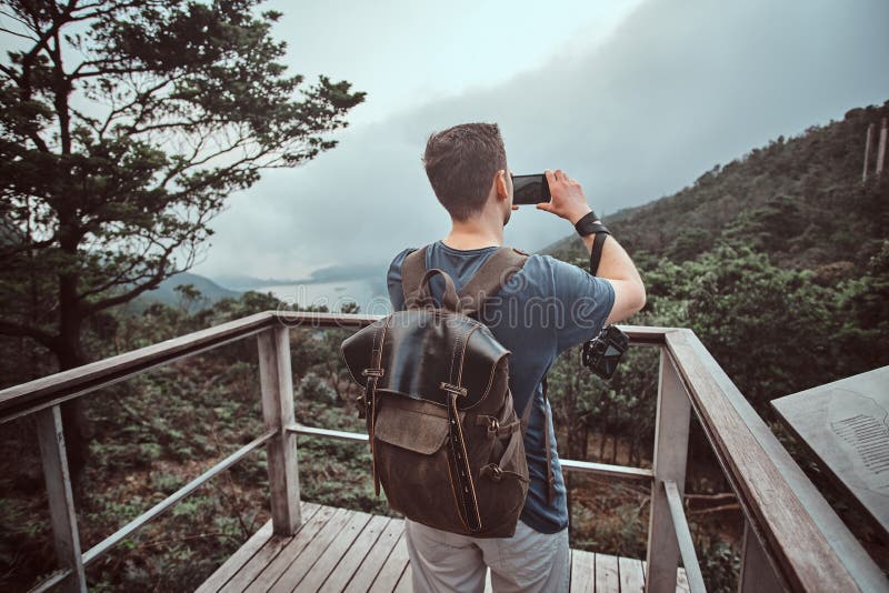 Young man with backpack is taking photo of beautiful nature while standing on sightseeing balcony