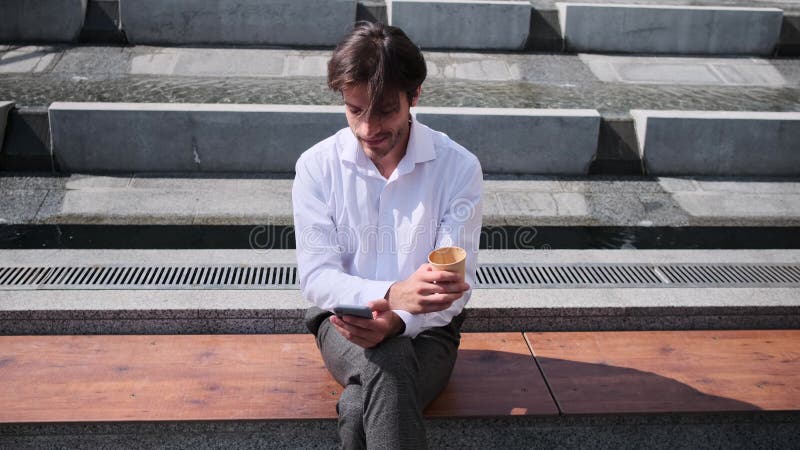 Young man drinking coffee and using mobile phone sitting on bench stock footage