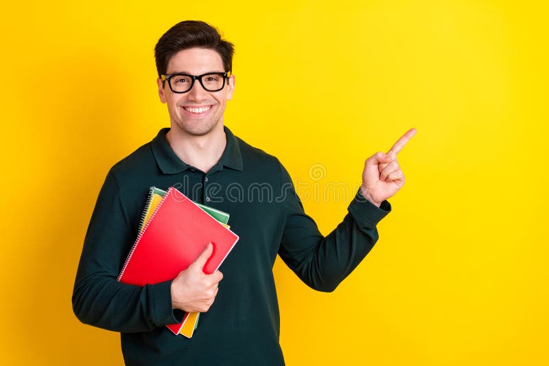Young man in green polo holding notebooks while pointing to the side against a vibrant yellow background, smiling royalty free stock image