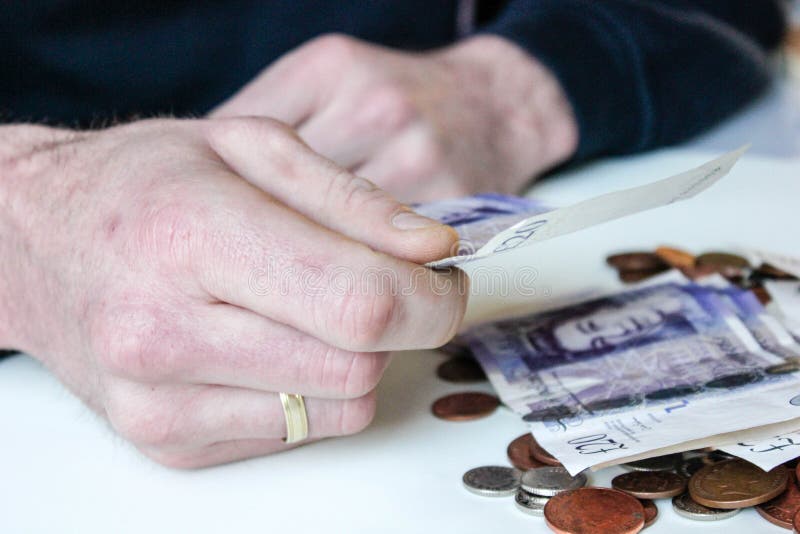 Young man holds a GBP twenty pounds money note and coins are liyng on a table around