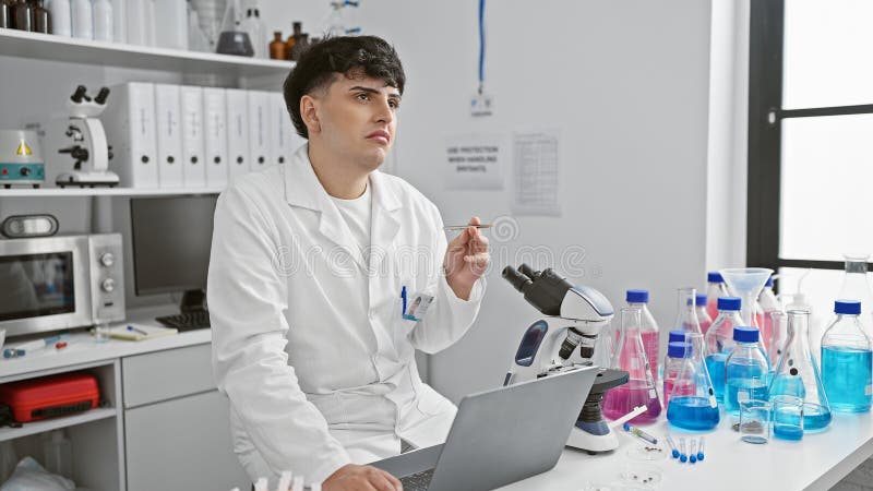 Young man in lab coat pondering a test tube in a modern laboratory with microscope and laptop royalty free stock photography