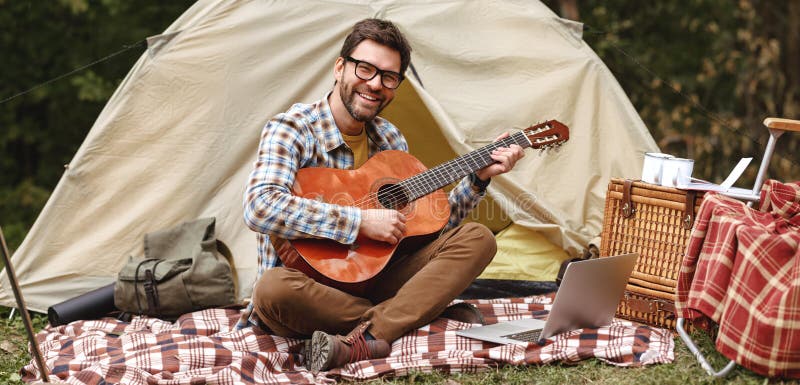 Young man learning to play acoustic guitar through internet on laptop during camping in forest