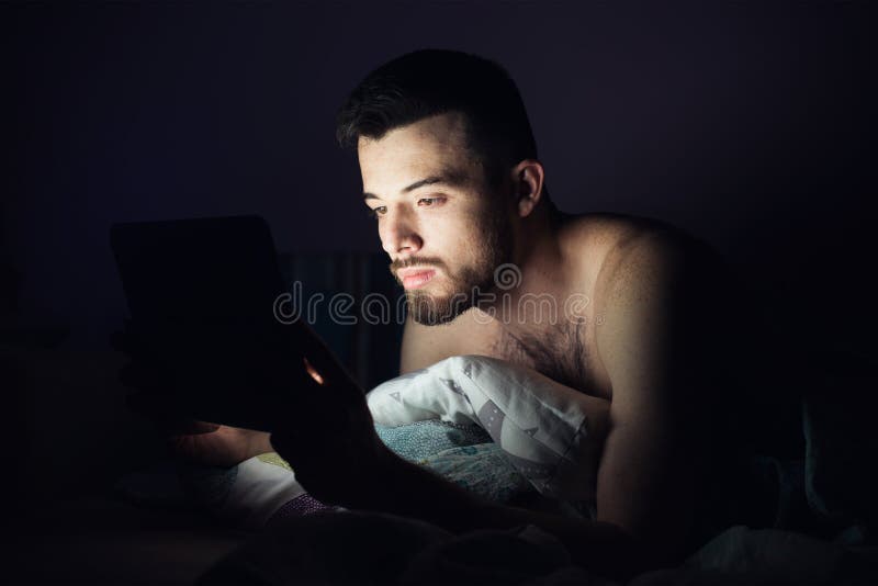 Young man lying on bed on his belly and look at tablet`s screen. Concentrated calm guy working playing games or