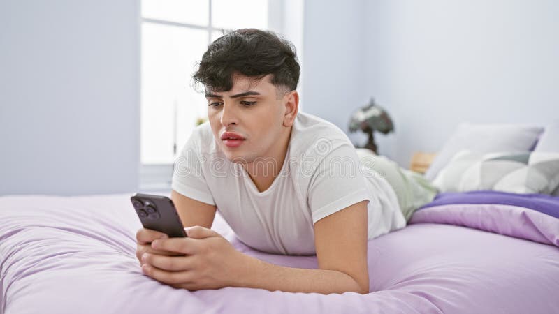 A young man lying on a purple bed using a smartphone, looking pensive in a bright bedroom royalty free stock photo