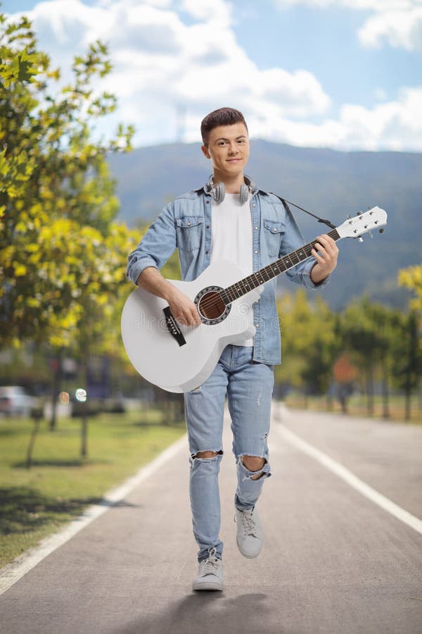 Young man playing an acoustic guitar and walking outdoors on a pedestiran lane