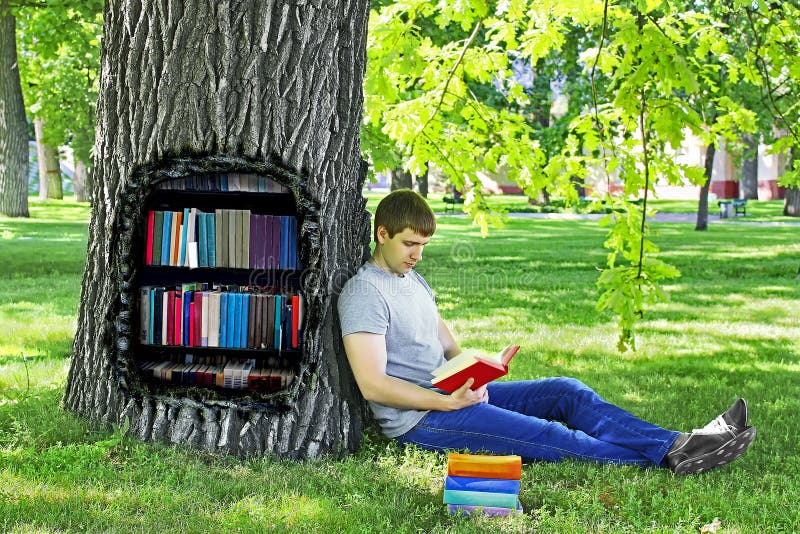 Young man reading a book sitting on the green grass leaning on a tree in the Park.