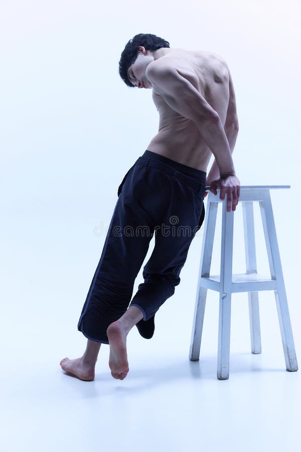 Young man with relief, beautiful body, posing shirtless in black trousers on high chair against white studio background. Back view. Male body aesthetics, style, fashion, health, men& x27;s beauty concept