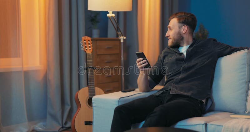 A young man sits contentedly on the couch in the living room in the evening