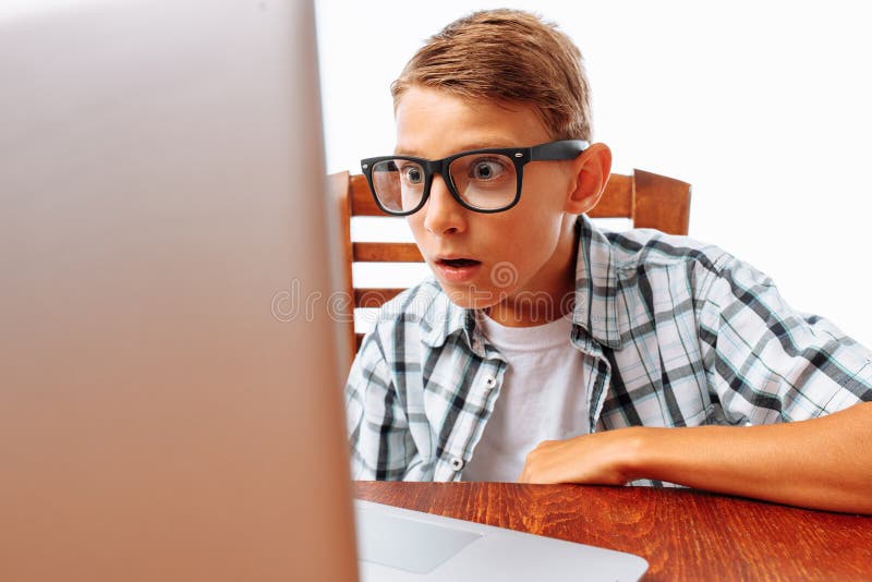 A young man sitting at a table with a laptop, shocked by what he saw, teen surprised look at the laptop, on a white background in