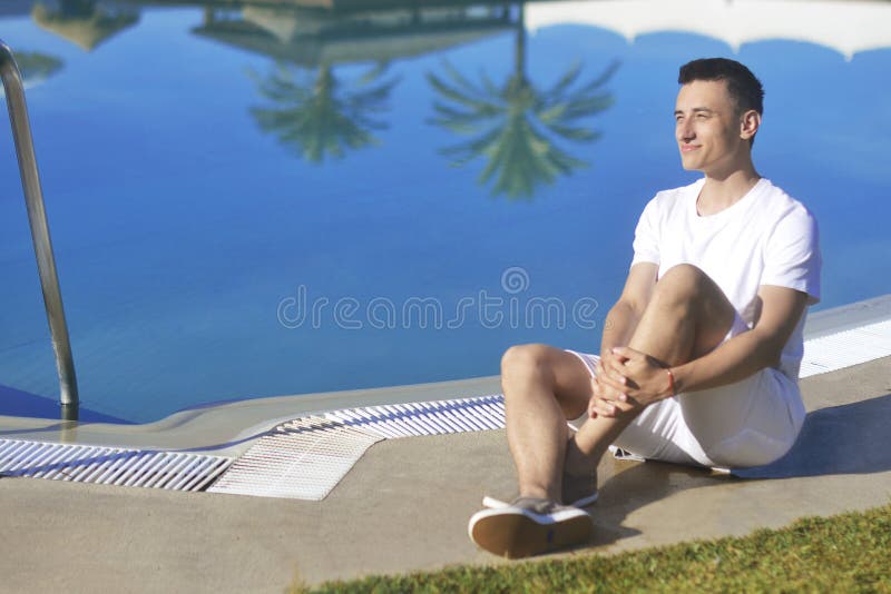 Young man smile in white clothes, posing near pool beackground palms. Man burns, relaxes, rests, travels. Sunbathing stock photos Young man smile in white clothes, posing near pool beackground palms. Man burns, relaxes, rests, travels. Sunbathing stock photos