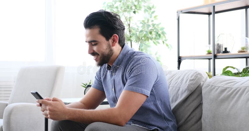 Young man smiling while using mobile phone sitting on sofa stock footage