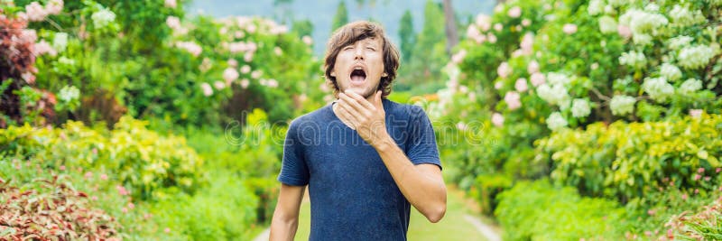 Young man sneeze in the park against the background of a flowering tree. Allergy to pollen concept BANNER long format