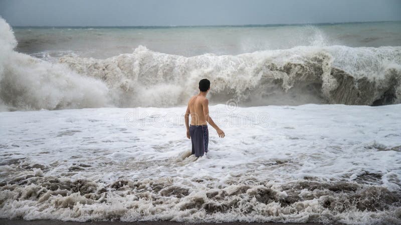 Young man standing against the sea waves with splash in a cloudy storm weather